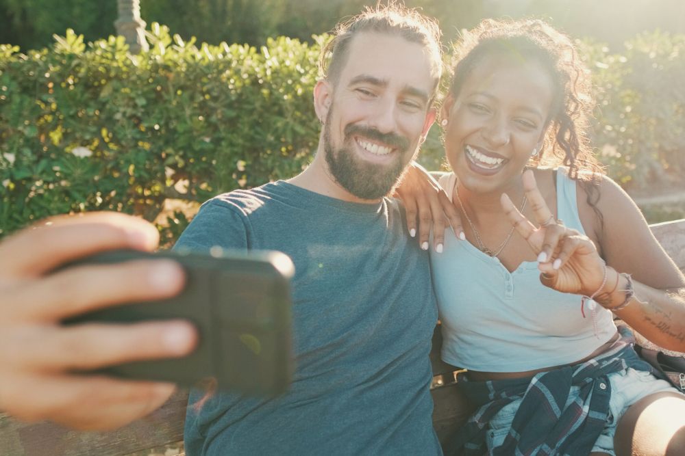 Happy smiling interracial couple taking selfie on phone while sitting on park bench Backlight