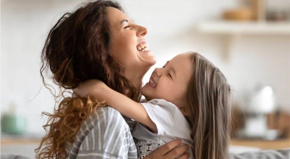 A mother and daughter embrace, laughing joyfully. They are in a bright, cozy indoor space with blurred kitchen elements in the background, conveying warmth and happiness.