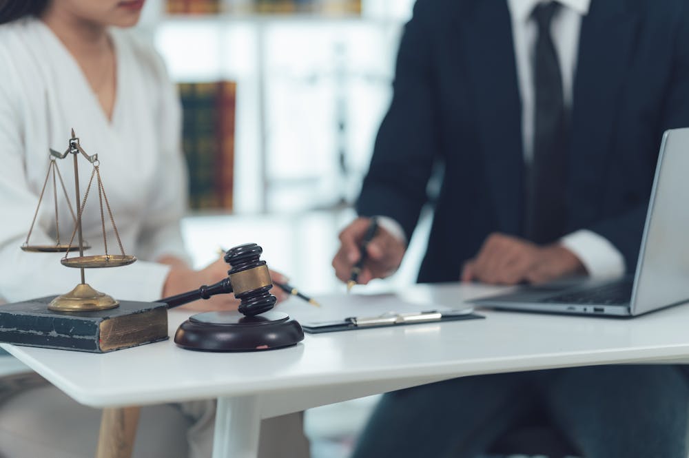 A gavel and scales of justice rest on a table as two professionals engage in discussion, with one writing notes beside a laptop in a well-organized office environment.